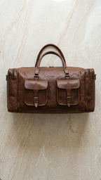 Brown Cedartrail Travel Duffel against pale stone, showing the twin handles, two buckled front pockets and zipped end compartments.
